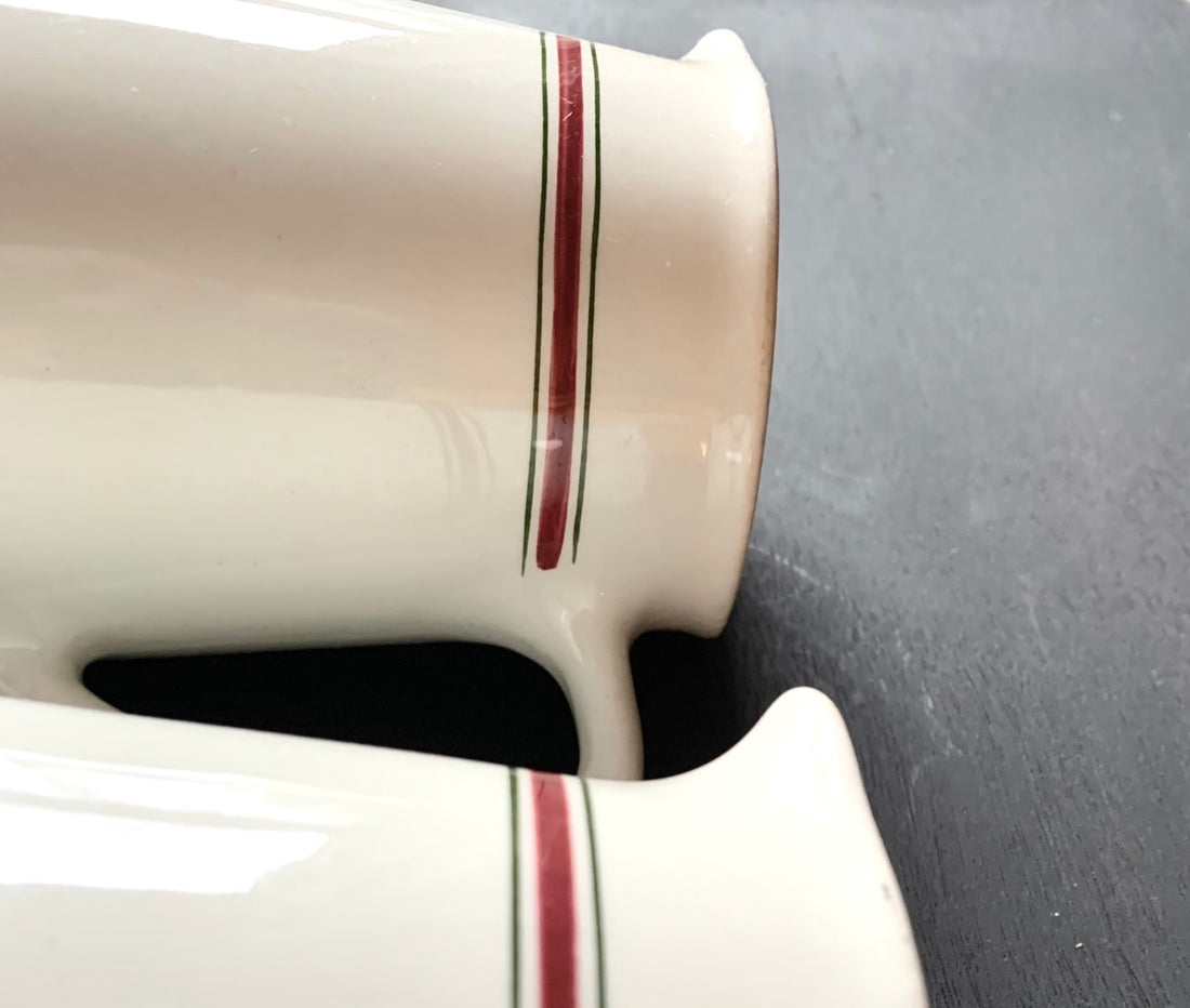 A close-up of the Pair English Stripe Pitchers, featuring white ceramic with red and black horizontal stripes near the edges, placed on a dark gray surface, evokes classic kitchen decor reminiscent of vintage milk pitchers.