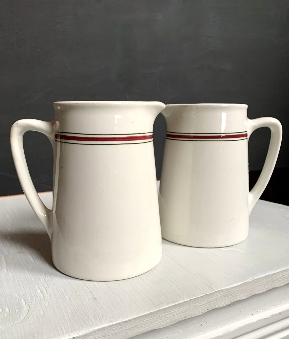 The Pair English Stripe Pitchers feature vintage red and black striped design, set on a white surface against a dark gray background, capturing the timeless appeal of Hercules vitrified pottery perfect for any kitchen decor.