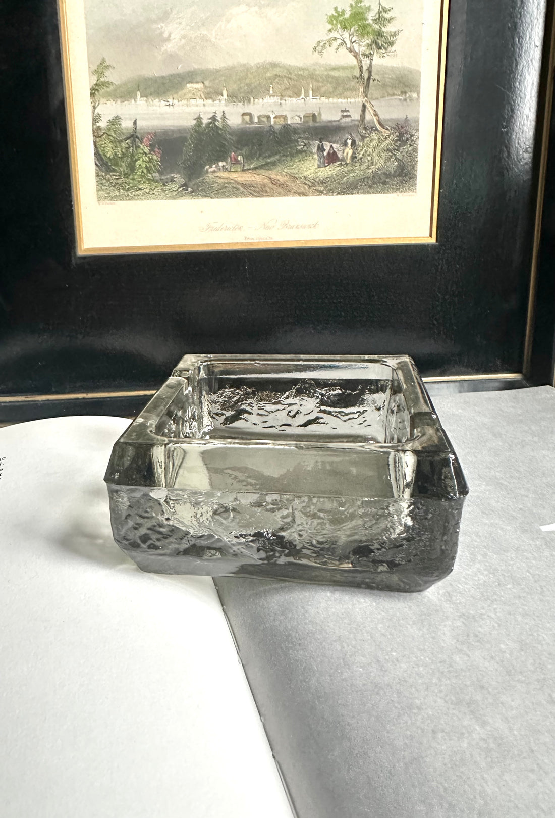 A square transparent green ashtray sitting on an open book with a black and white print in the background