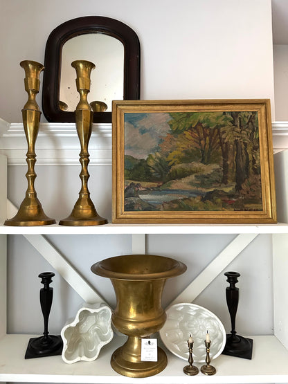 Decorative shelf with brass candlesticks, urn, and framed painting against a white wall.