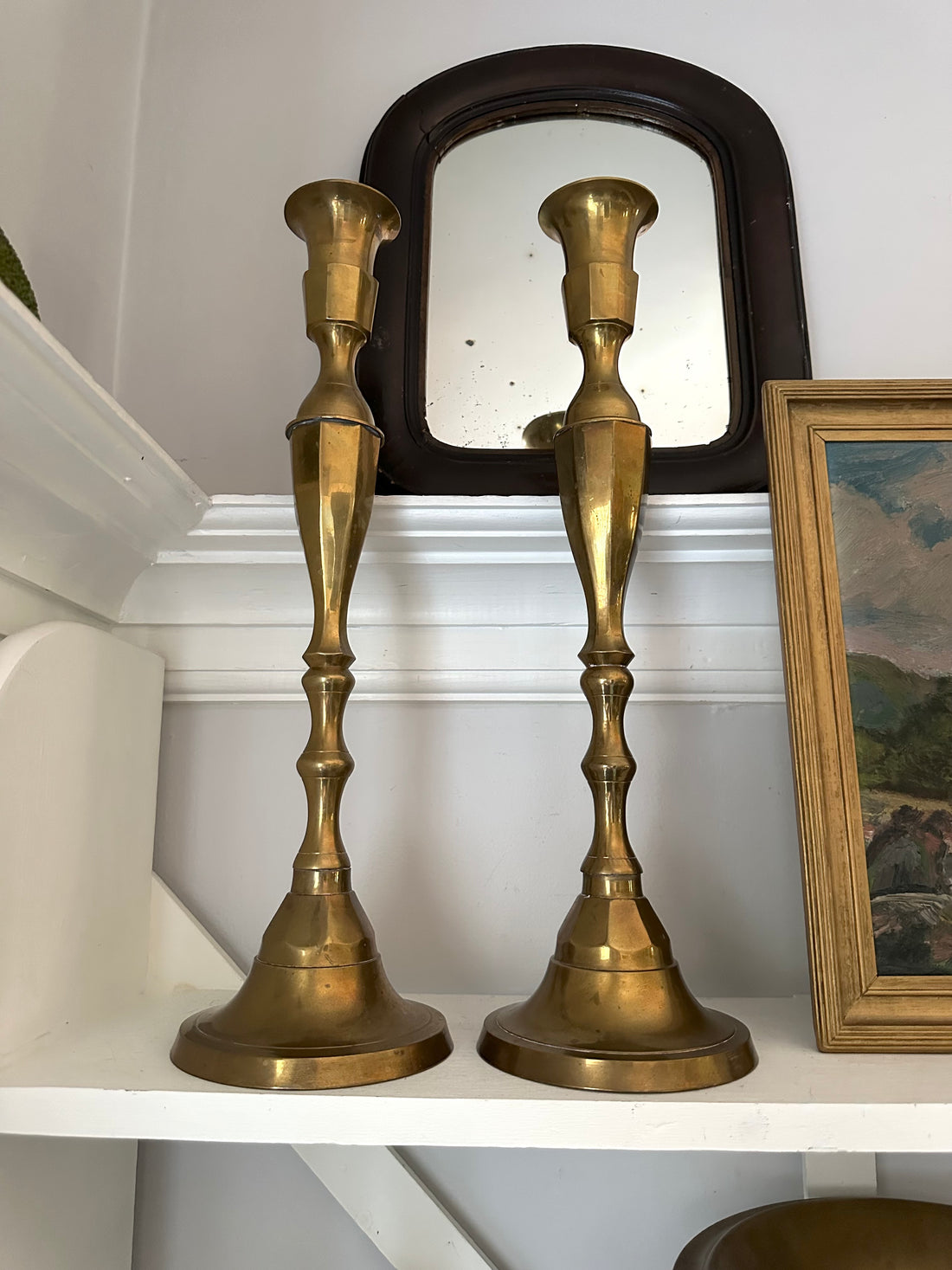 Two brass candlesticks on a shelf with a mirror and framed picture in the background.