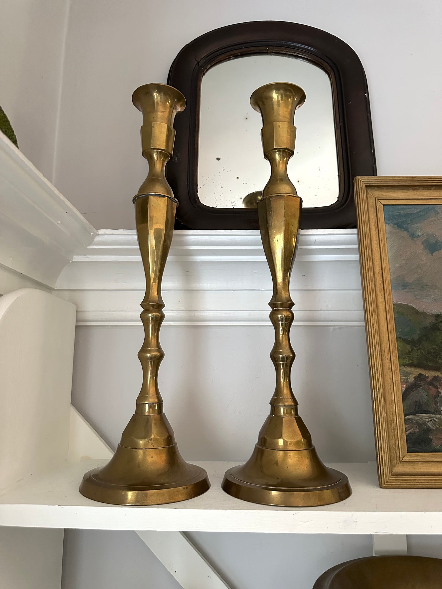 Two brass candlesticks on a shelf with a mirror and framed picture in the background.