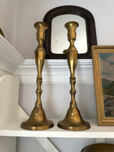 Two brass candlesticks on a shelf with a mirror and framed picture in the background.