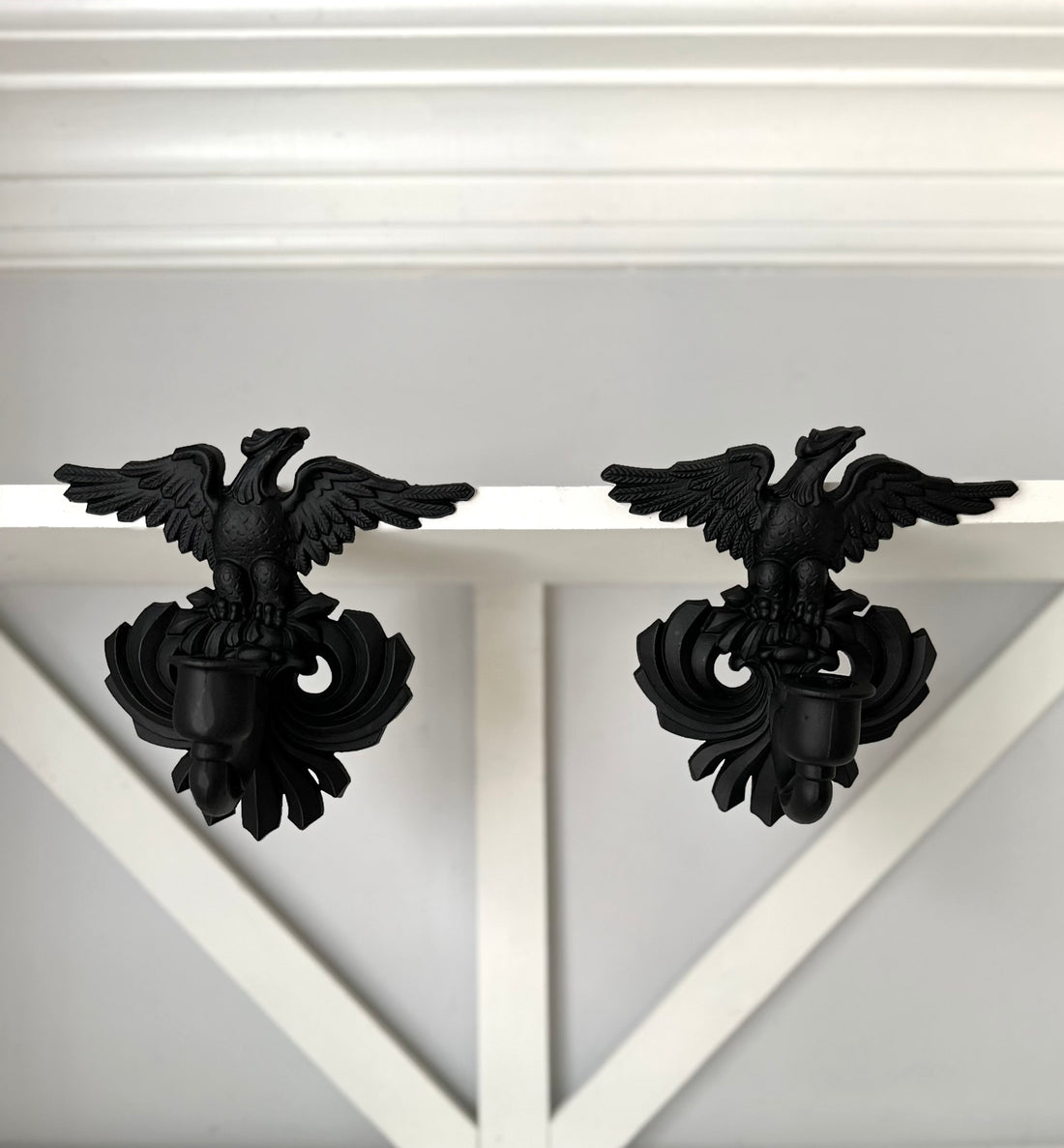 A pair of black metal eagle wall hooks mounted on a white wooden shelf against a light gray wall.