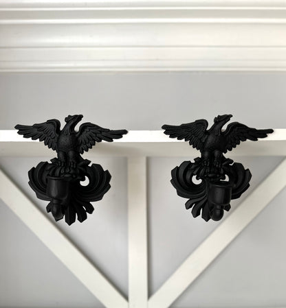 A pair of black metal eagle wall hooks mounted on a white wooden shelf against a light gray wall.