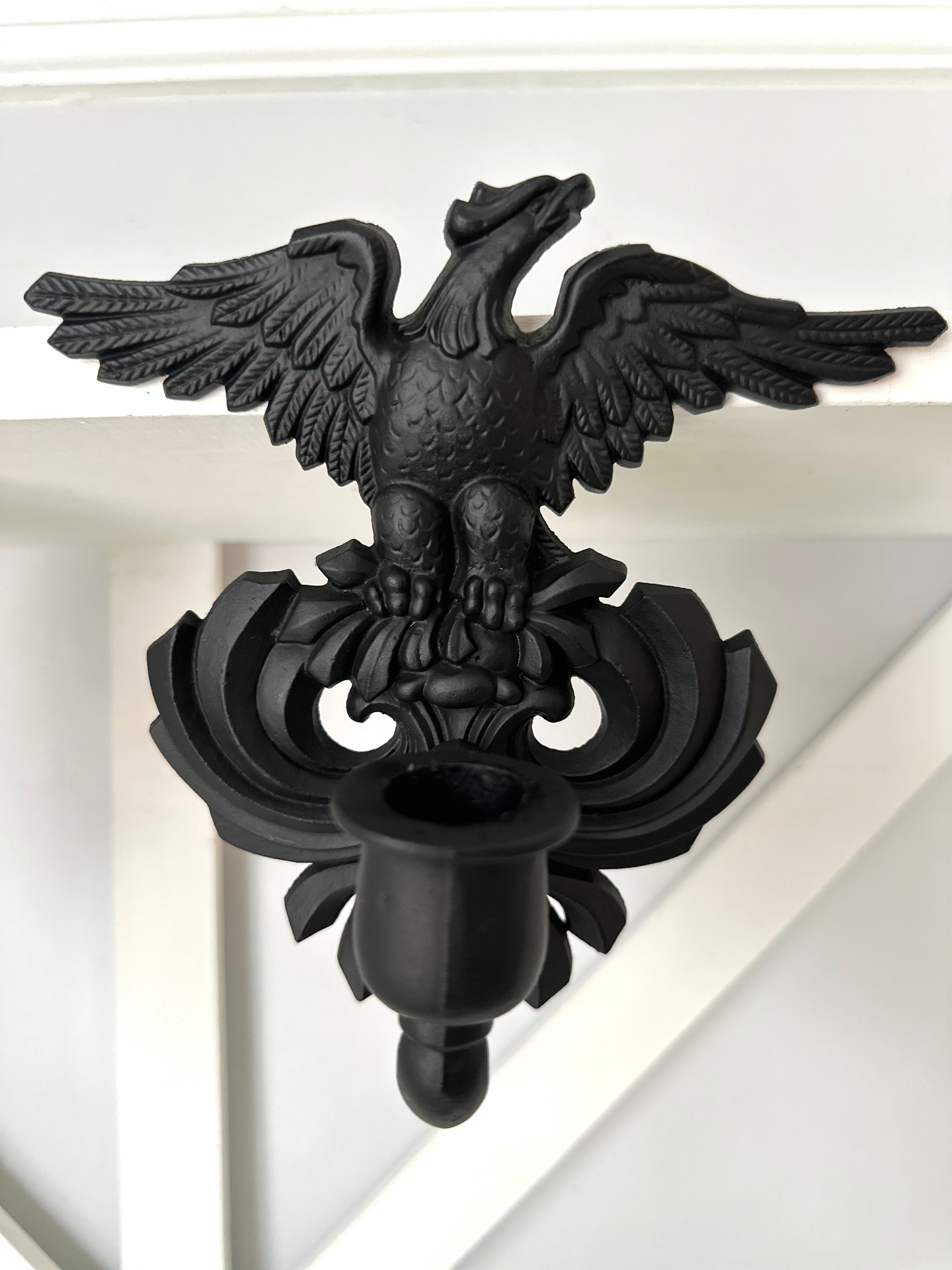 A black metal double eagle wall sconce mounted on a white painted wooden wall or shelf.