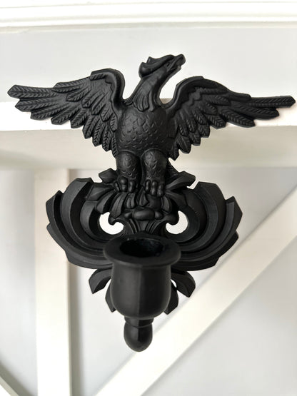 A black metal double eagle wall sconce mounted on a white painted wooden wall or shelf.