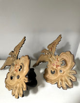 A pair of vintage-style wooden bird-shaped ornaments or decorative pieces with carved facial features and small red painted markings, displayed against a plain light background.