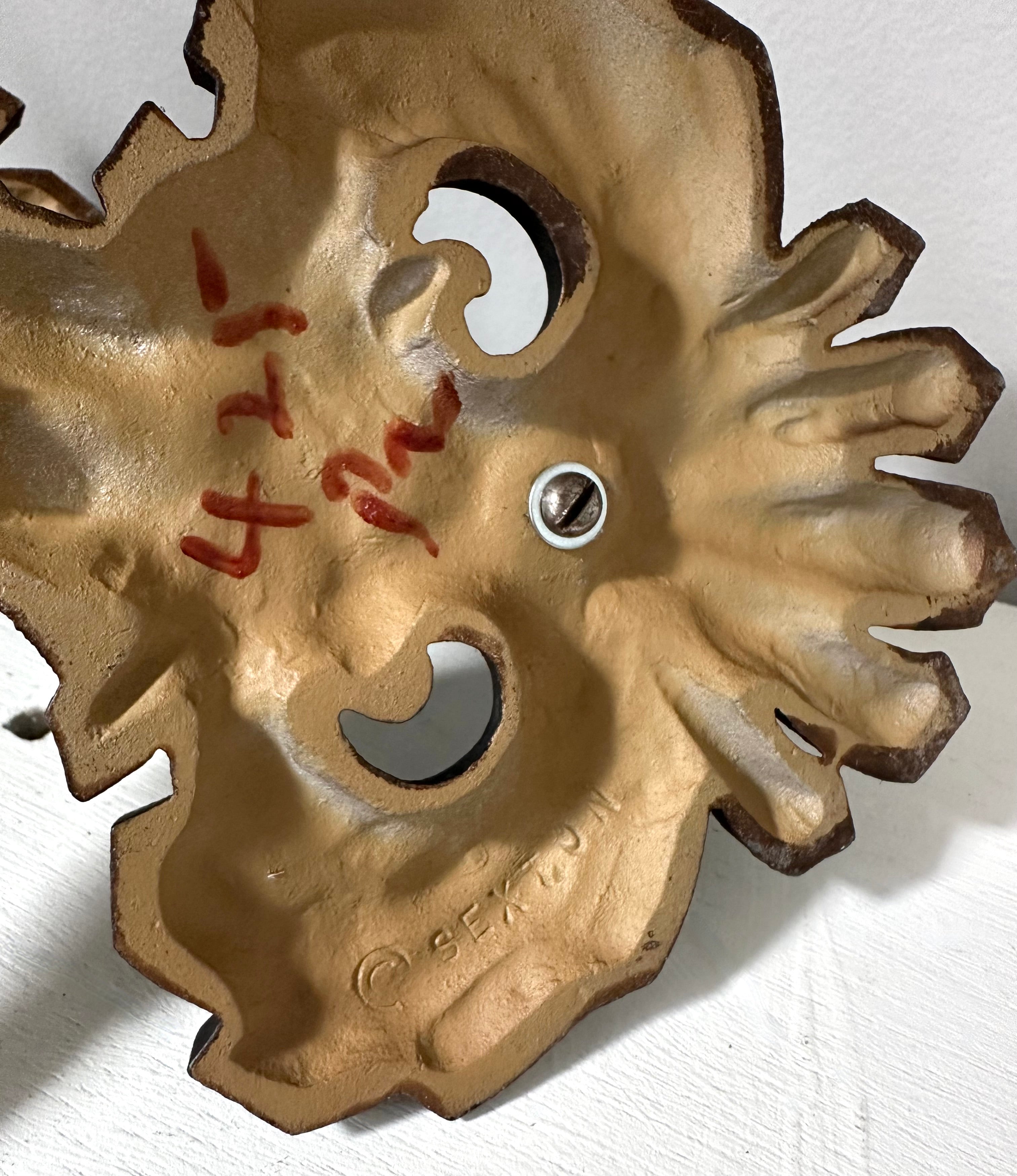 A close-up of a rustic, hand-painted wooden decorative piece with a weathered tan-brown finish, featuring carved details, a small silver screw, and red handwritten markings on a white textured background.