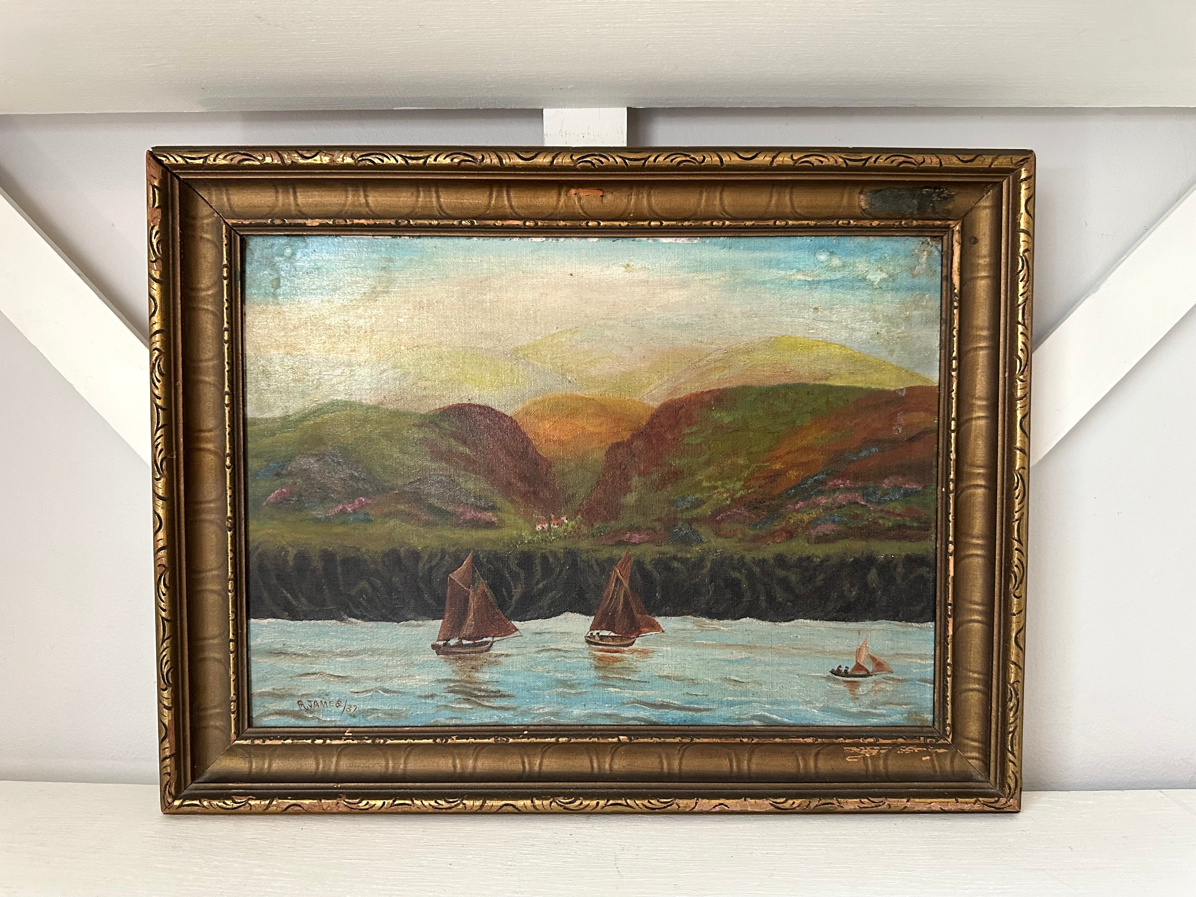 An oil painting of a coastal landscape with sailboats on the water, framed in an ornate gold-toned frame and displayed against a white wall.