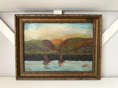 An oil painting of a coastal landscape with sailboats on the water, framed in an ornate gold-toned frame and displayed against a white wall.