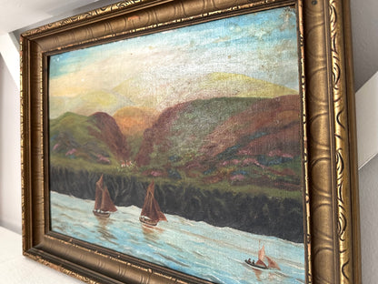 An oil painting of a coastal landscape with sailboats on the water, framed in an ornate gold frame and shown hanging on a white wall.