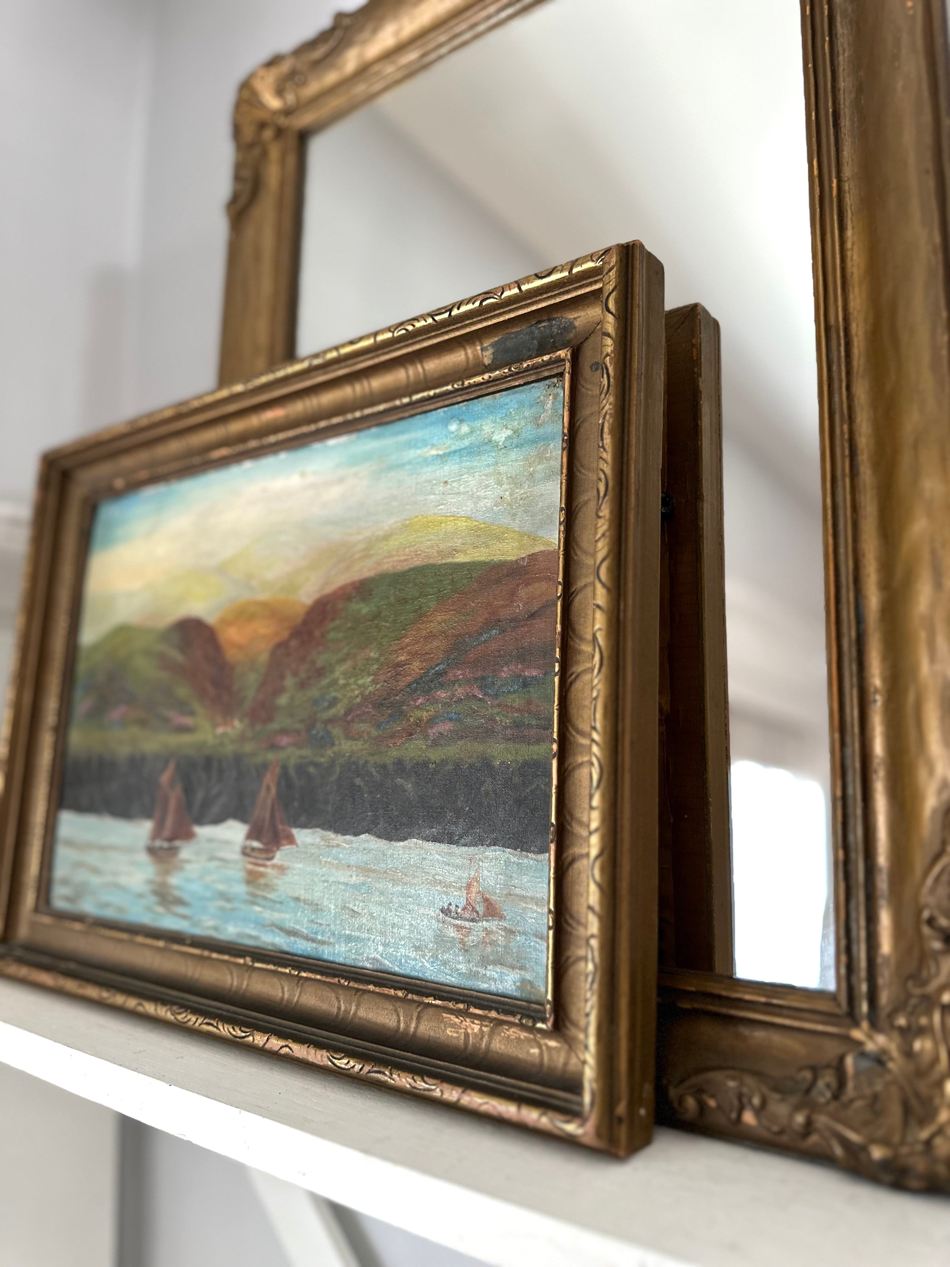 An antique framed landscape oil painting with a detailed gold frame, leaning on a white shelf next to another ornate frame.
