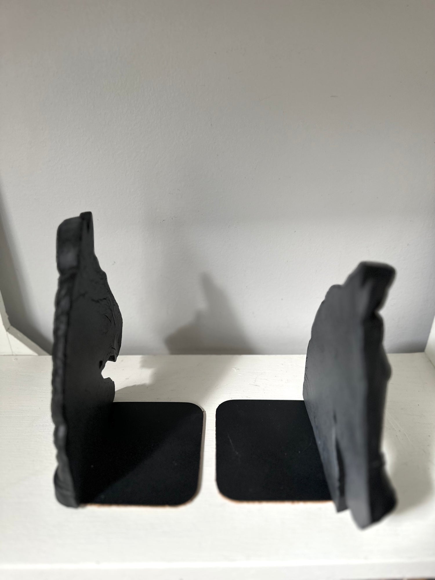 A pair of black bookends shaped like stylized animal heads, standing on square bases on a white shelf against a plain light gray wall.