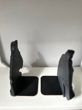 A pair of black bookends shaped like stylized animal heads, standing on square bases on a white shelf against a plain light gray wall.