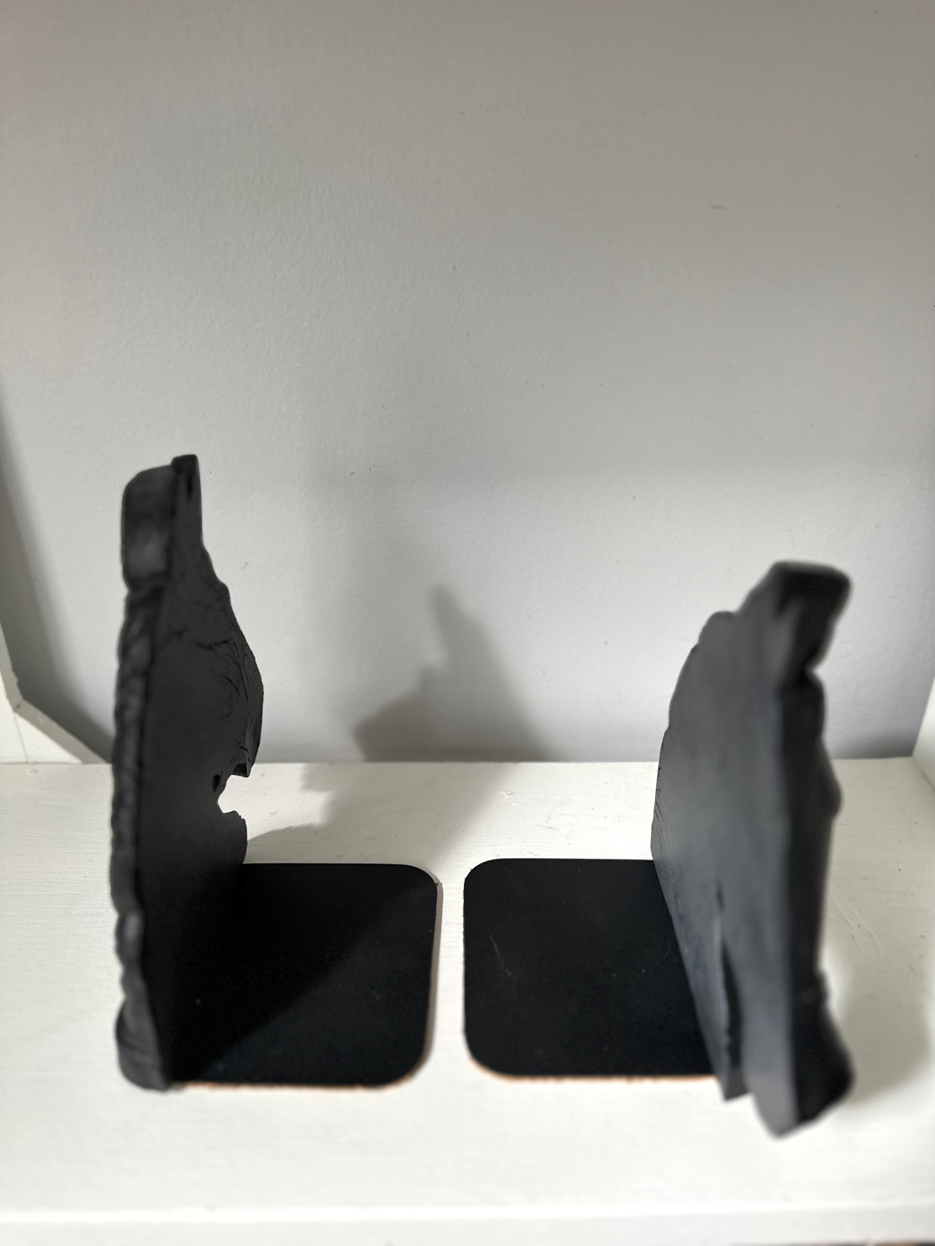 A pair of black bookends shaped like stylized animal heads, standing on square bases on a white shelf against a plain light gray wall.