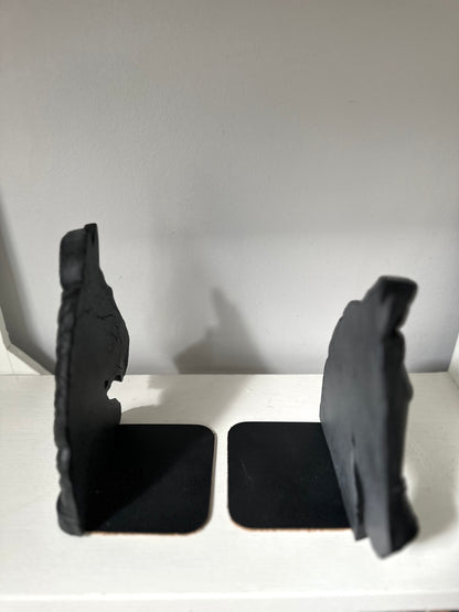 A pair of black bookends shaped like stylized animal heads, standing on square bases on a white shelf against a plain light gray wall.
