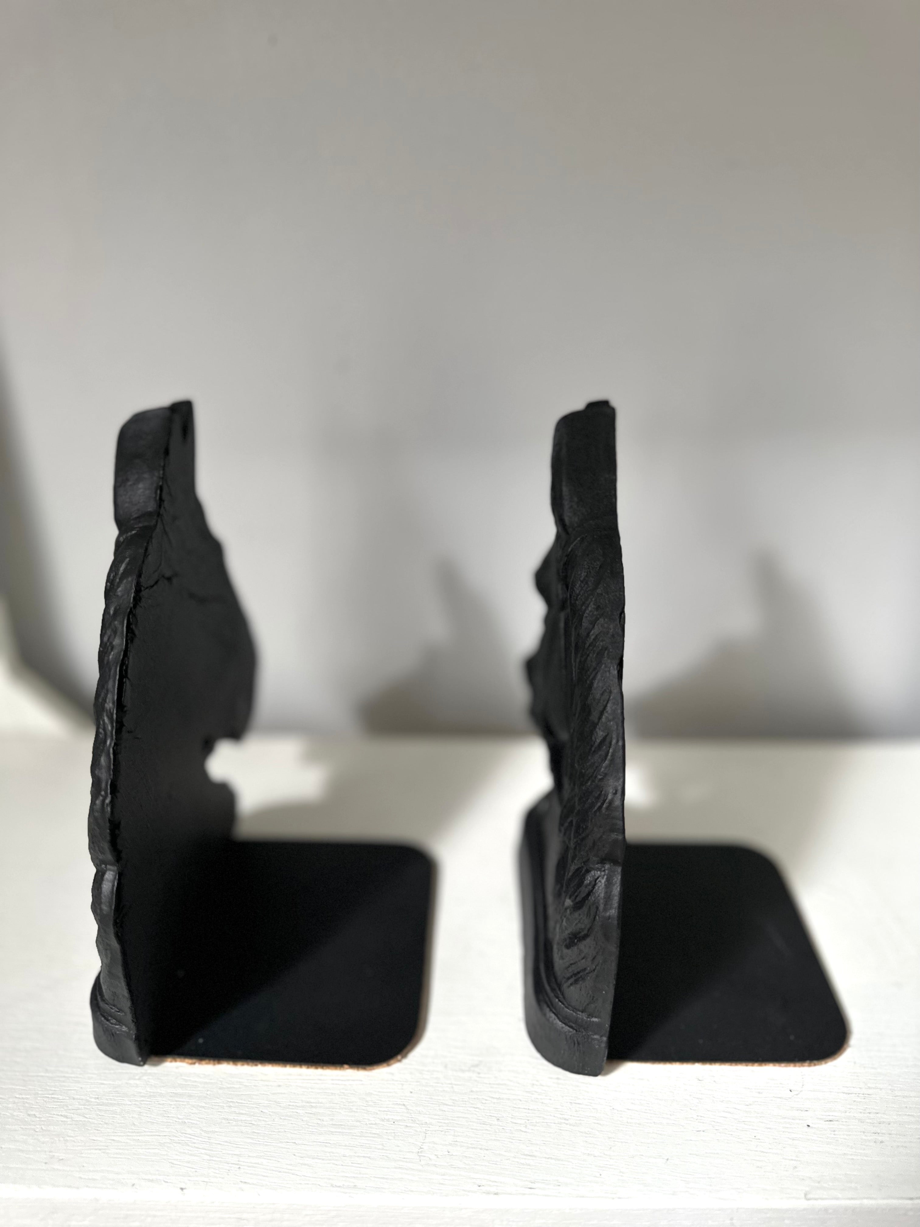 A pair of black rustic stone bookends with a carved mountain or cliff face design, standing on a white surface against a light gray background.