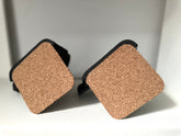A pair of square cork coasters with black rubber borders, shown standing upright on a white surface against a light gray background.