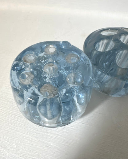 A pair of round, translucent light blue glass candleholders with multiple circular cutouts, displayed on a white textured surface.