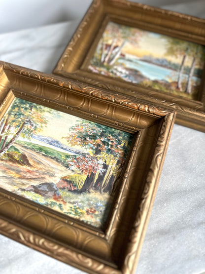 Two small framed landscape paintings with ornate gold frames, resting on a white fabric surface.