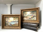 Two framed landscape paintings with ornate gold frames, displayed on a white shelf with dark books underneath the right frame.