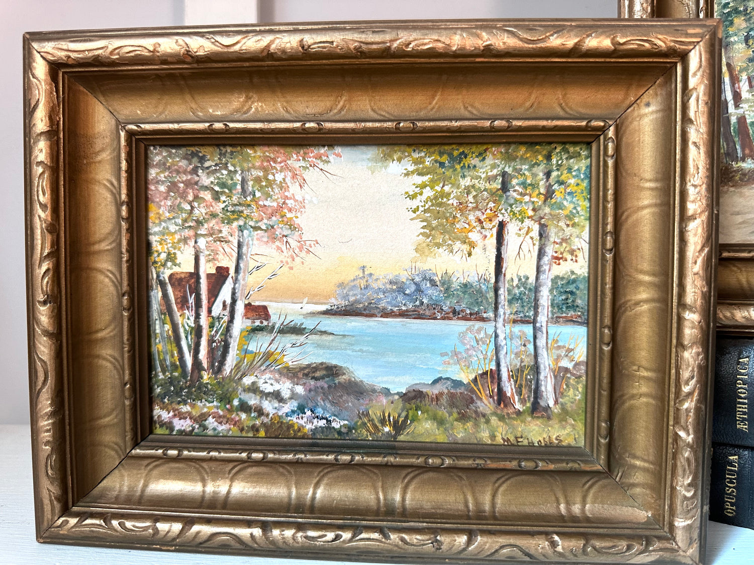 A framed landscape oil painting featuring a lake, trees, and shoreline, housed in an ornate gold-toned carved frame. The painting is displayed leaning against other framed art on a light surface.