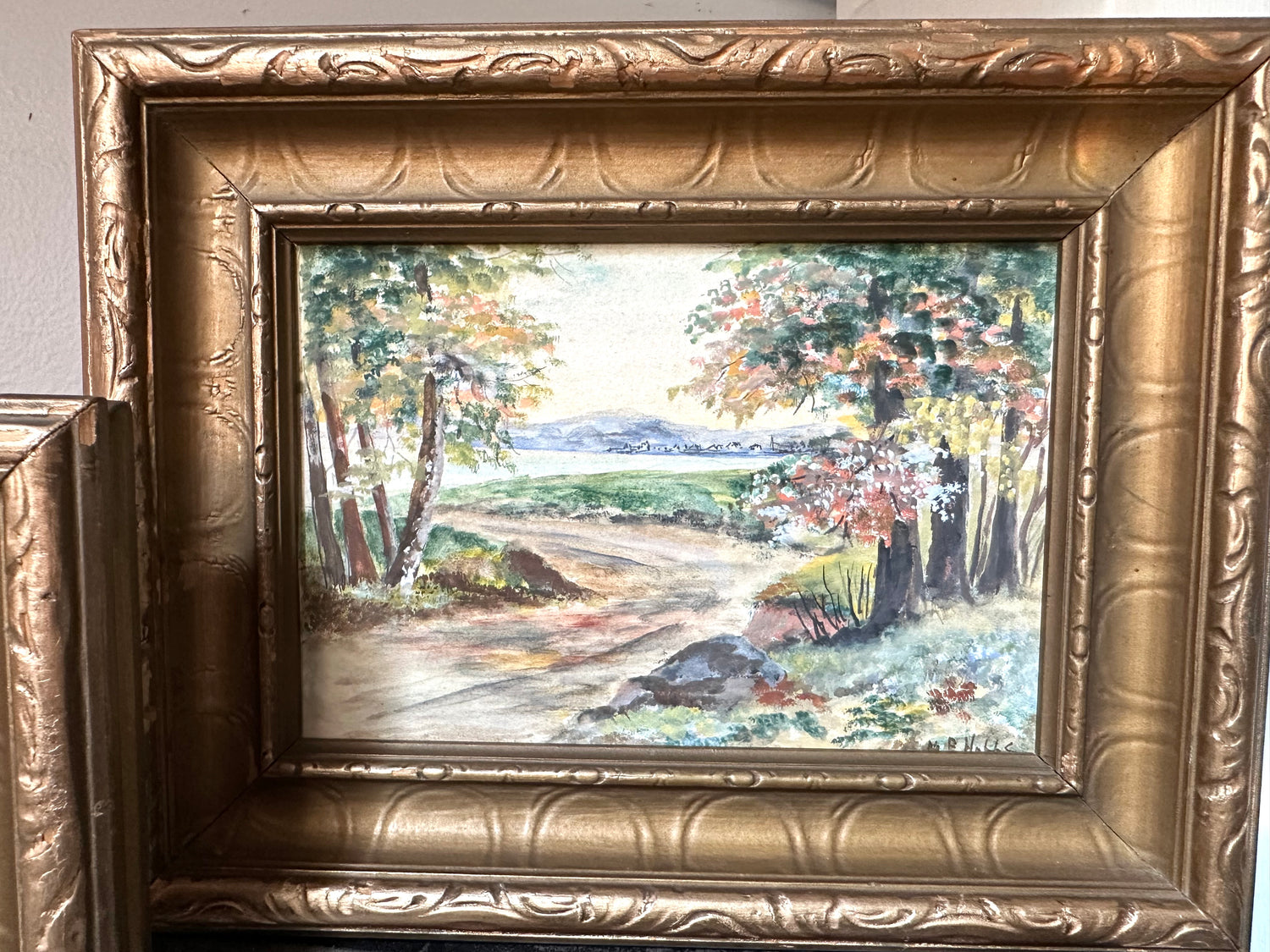 A framed landscape painting showing a dirt road winding through trees with green, yellow, and brown foliage, set in an ornate gold frame.