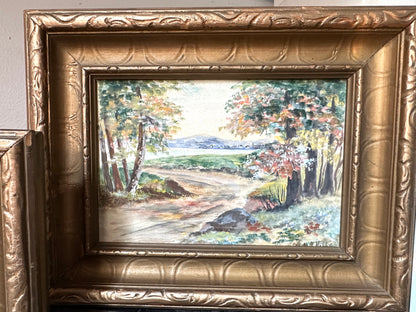 A framed landscape painting showing a dirt road winding through trees with green, yellow, and brown foliage, set in an ornate gold frame.